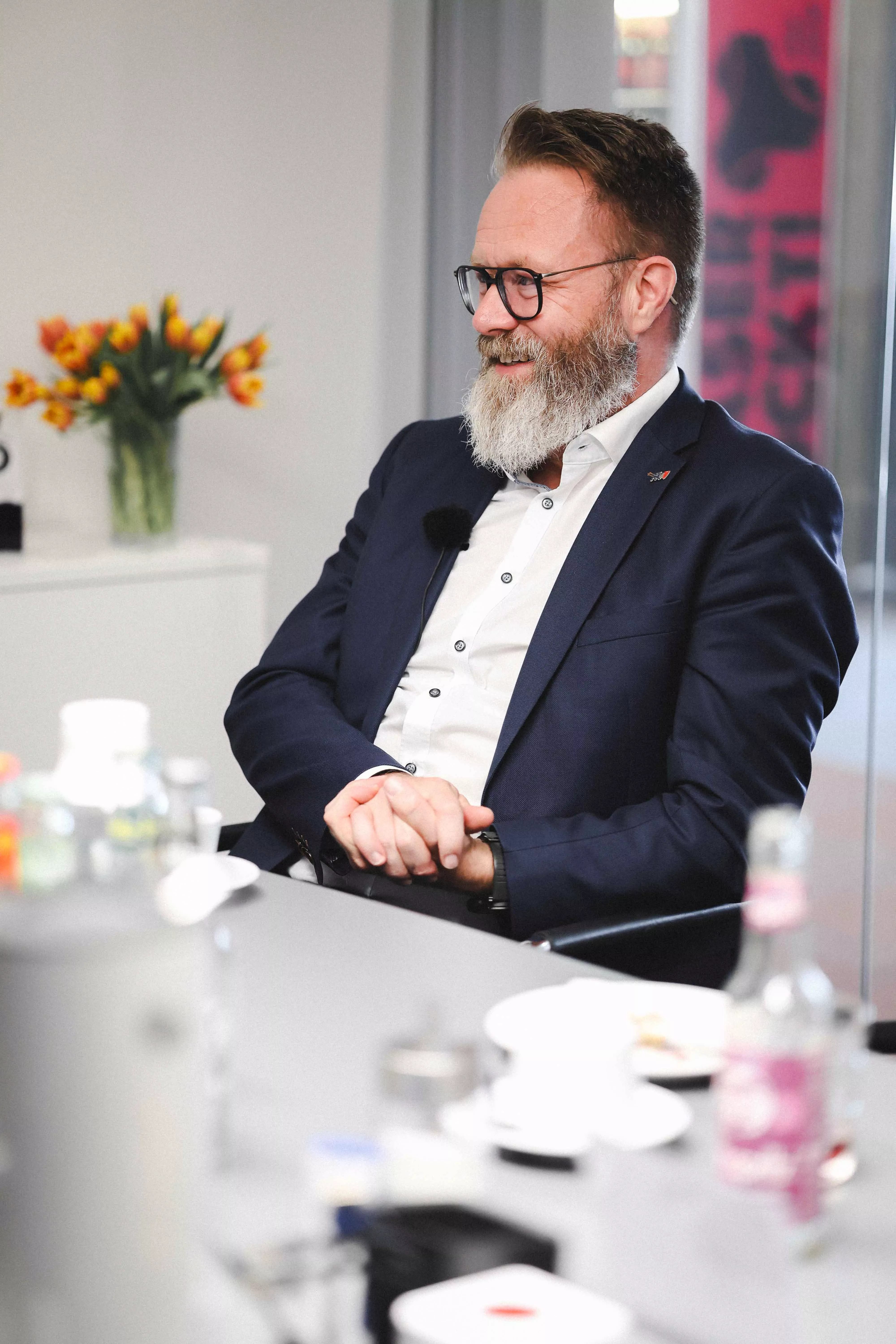 Claus Ruhe Madsen hört aufmerksam lächelnd zu und hat die Hände ineinander verschränkt. | Claus Ruhe Madsen listening attentively with a smile, hands folded.
