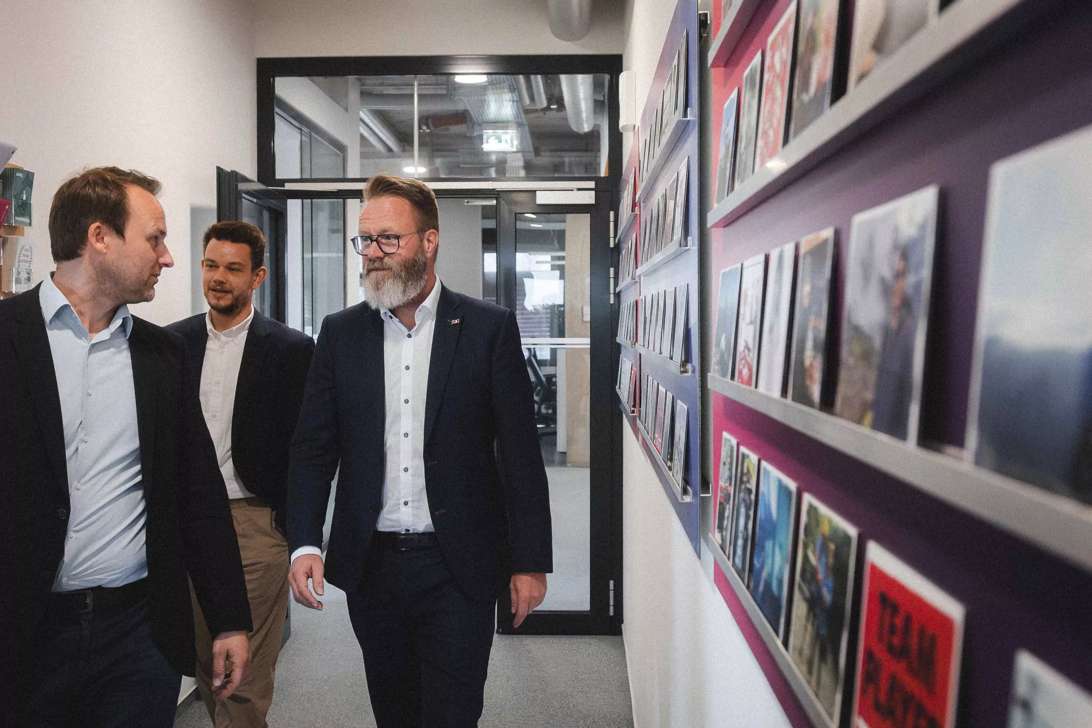 Johannes Ripken, Alexander Ubben und Claus Ruhe Madsen gehen im TRASER-Büro den Flur entlang an der Fotowand. | Johannes Ripken, Alexander Ubben and Claus Ruhe Madsen walk down the hallway past the photo wall at the TRASER office.