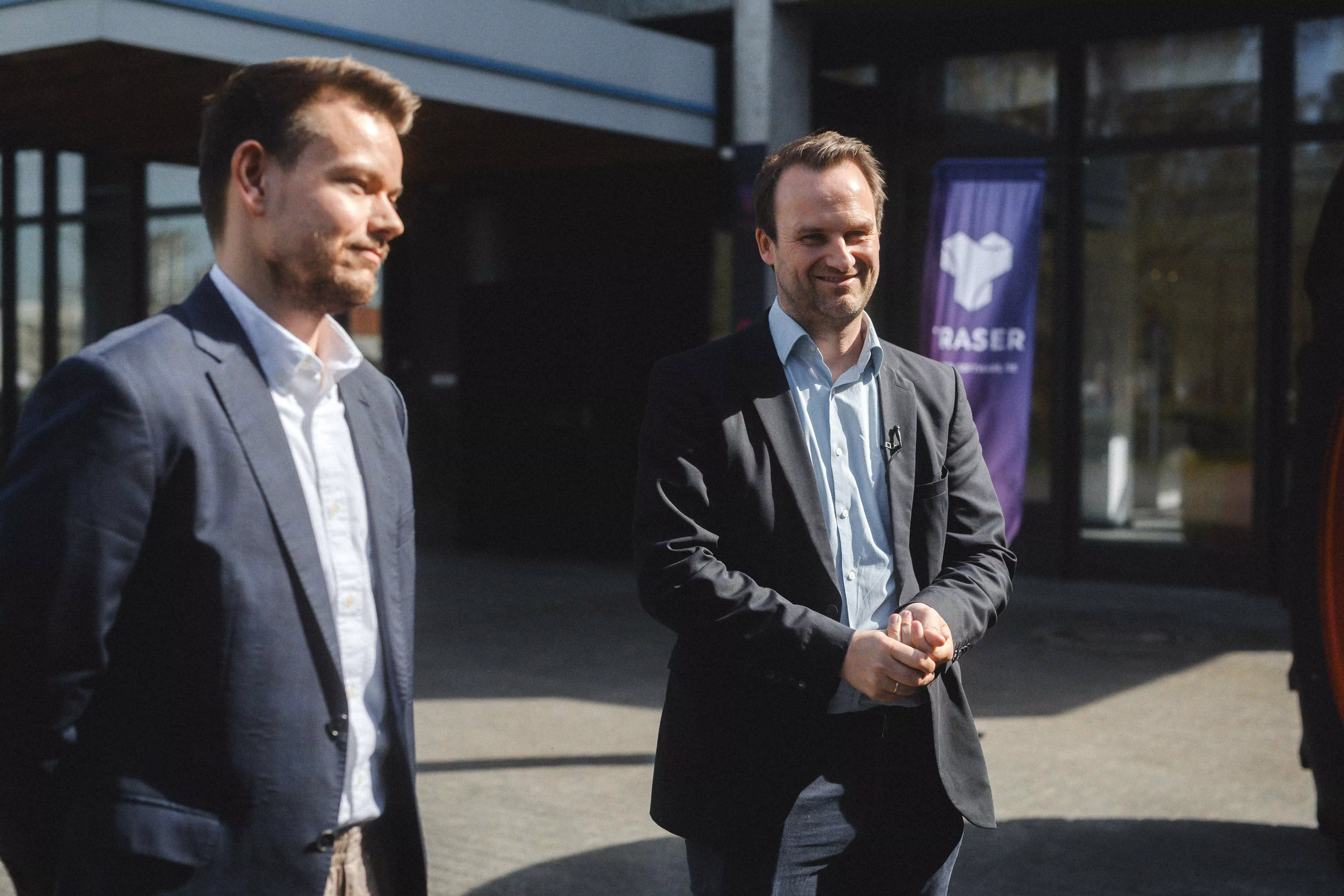Draußen vor dem Bürogebäude: Johannes Ripken im Fokus, Alexander Ubben seitlich und unscharf im Vordergrund; im Hintergrund eine TRASER Beachflag. | Outside the office building: Johannes Ripken in focus, Alexander Ubben seen from the side and blurred in the foreground; a TRASER beach flag in the background.